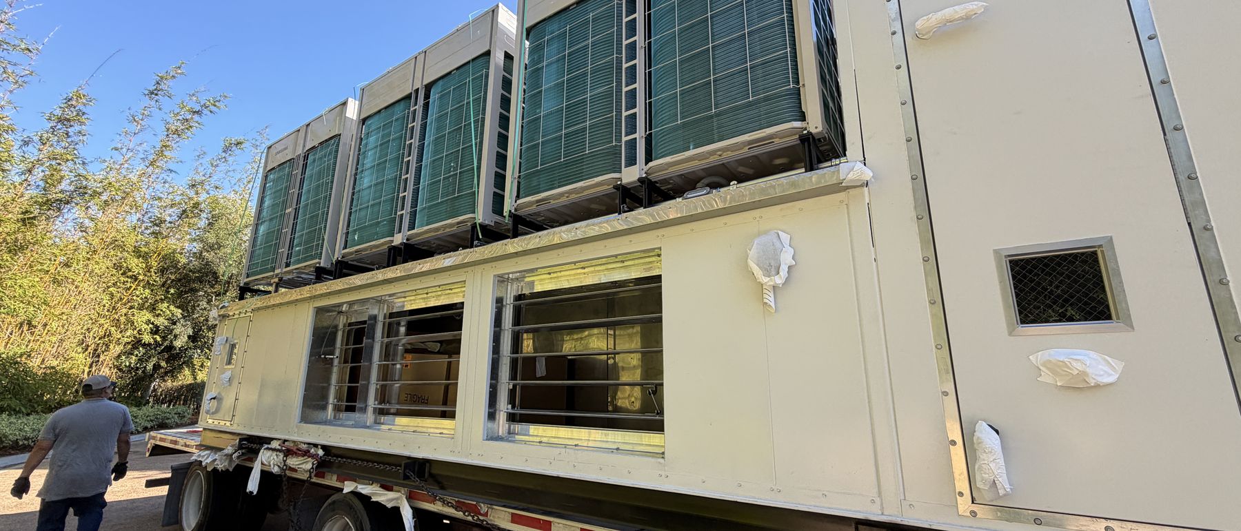 Large packaged rooftop unit arriving by flatbed alongside a multi-storey office building